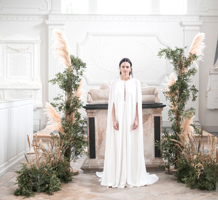 Bride in Jesus Peiro Wedding Dress and Bridal Cape Standing in Front of a Floral installation of Pampas Grass and Dried Flowers