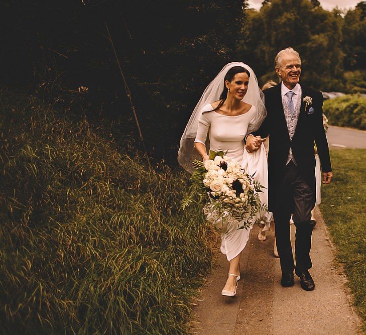 Bride walking to the church with her father clutching a beautiful white rose bouquet