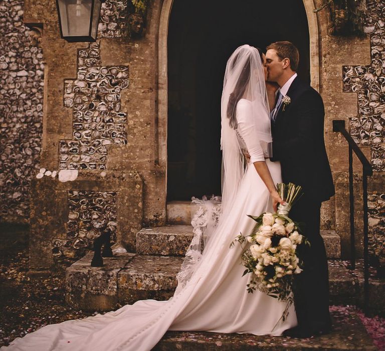 Bride and groom kiss at the church doorway with beautiful floral arch and white rose bouquet