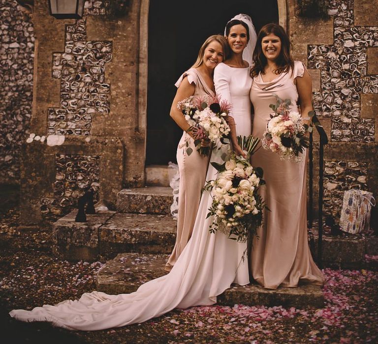 Bridesmaids wearing simple oyster dresses with bride clutching white rose bouquets