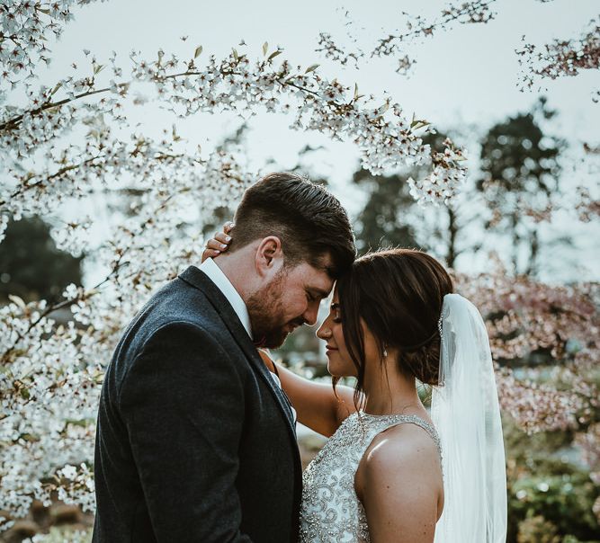 Bride in Halterneck Wedding Dress and Groom in Dark Suit Embracing by Blossom Trees