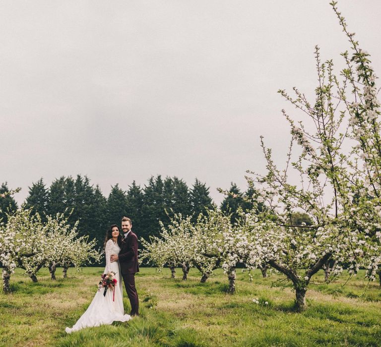 Groom in Burgundy Suit and Bride in Lace Wedding Dress Standing in an Orchard