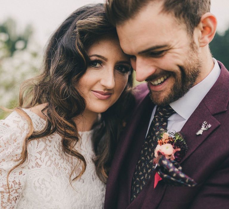 Groom in Burgundy Suit and Bride in Lace Wedding Dress Embracing in an Orchard