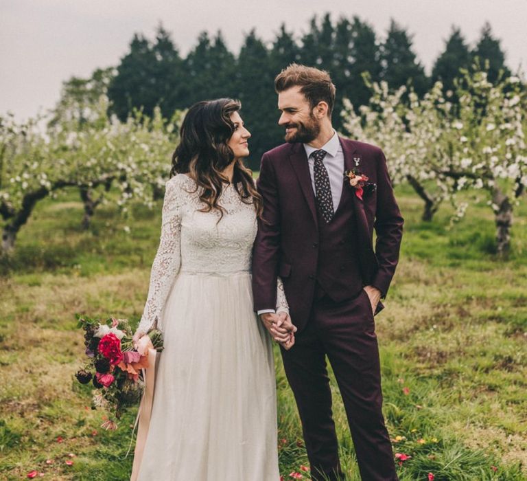 Bride in Lace Wedding Dress and Groom in Burgundy Suit Holding Hands in an Orchard