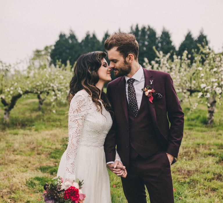 Bride in Lace Wedding Dress and Groom in Burgundy Suit Kissing in an Orchard