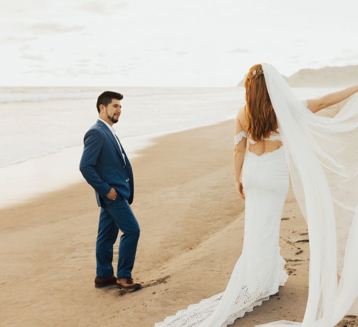 Bride Holds Up Veil On Beach With Groom In Navy Suit