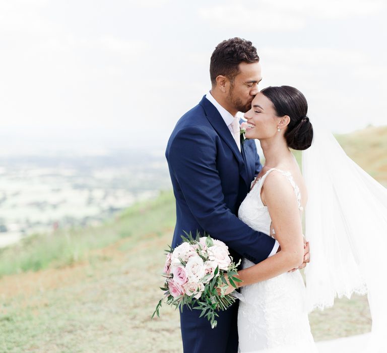 Groom in blue suit kissing his brides head in a Pronovias wedding dress