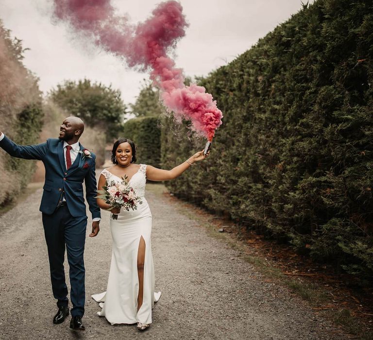 Groom in navy suit and bride in wedding dress with front split holding coloured smoke flares