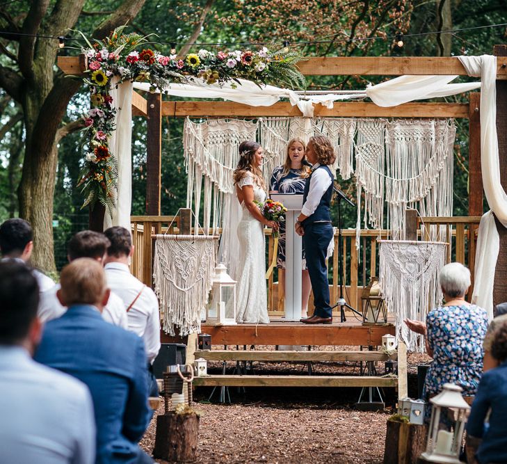 Bride in Lenora Dress by Wtoo Watters with Lace Cap Sleeves and Keyhole Back | Groom in Blue Waistcoat and Trousers with Colourful Patterned Bow Tie |  Colourful Bridal Bouquet with Sunflowers and Yellow Trailing Ribbon | Wooden Altar Decorated with Macrame Hangings | Woodland Wedding Ceremony at Lila's Wood | Macrame Decor, Vintage Caravan Photobooth and Five-Tier Naked Wedding Cake for Boho Wedding in Woodlands | Freckle Photography