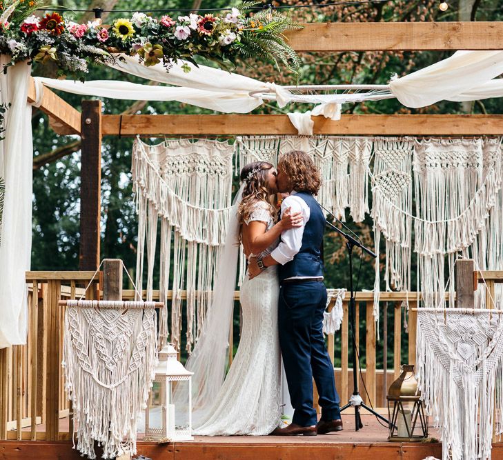 Bride in Lenora Dress by Wtoo Watters with Lace Cap Sleeves and Keyhole Back | Groom in Blue Waistcoat and Trousers with Colourful Patterned Bow Tie | Wooden Altar Decorated with Macrame Hangings | Woodland Wedding Ceremony at Lila's Wood | Macrame Decor, Vintage Caravan Photobooth and Five-Tier Naked Wedding Cake for Boho Wedding in Woodlands | Freckle Photography