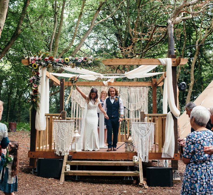 Bride in Lenora Dress by Wtoo Watters with Lace Cap Sleeves and Keyhole Back | Groom in Blue Waistcoat and Trousers with Colourful Patterned Bow Tie |  Colourful Bridal Bouquet with Sunflowers and Yellow Trailing Ribbon | Wooden Altar Decorated with Macrame Hangings | Woodland Wedding Ceremony at Lila's Wood | Macrame Decor, Vintage Caravan Photobooth and Five-Tier Naked Wedding Cake for Boho Wedding in Woodlands | Freckle Photography