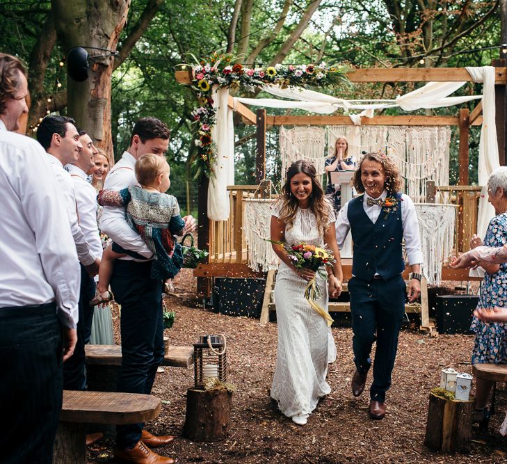 Bride in Lenora Dress by Wtoo Watters with Lace Cap Sleeves and Keyhole Back | Groom in Blue Waistcoat and Trousers with Colourful Patterned Bow Tie |  Colourful Bridal Bouquet with Sunflowers and Yellow Trailing Ribbon | Wooden Altar Decorated with Macrame Hangings | Woodland Wedding Ceremony at Lila's Wood | Macrame Decor, Vintage Caravan Photobooth and Five-Tier Naked Wedding Cake for Boho Wedding in Woodlands | Freckle Photography