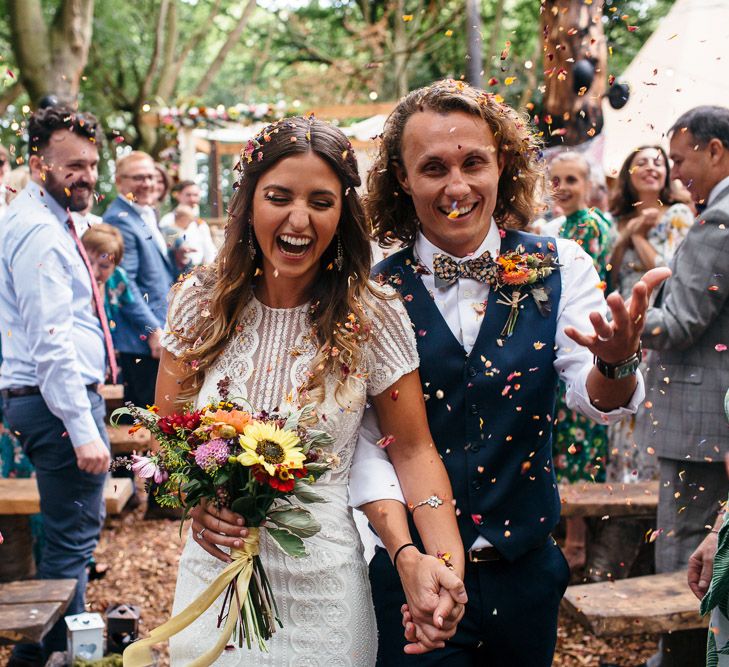 Bride in Lenora Dress by Wtoo Watters with Lace Cap Sleeves and Keyhole Back | Groom in Blue Waistcoat and Trousers with Colourful Patterned Bow Tie |  Colourful Bridal Bouquet with Sunflowers and Yellow Trailing Ribbon | Woodland Wedding Ceremony at Lila's Wood | Macrame Decor, Vintage Caravan Photobooth and Five-Tier Naked Wedding Cake for Boho Wedding in Woodlands | Freckle Photography