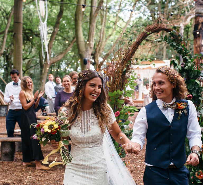 Bride in Lenora Dress by Wtoo Watters with Lace Cap Sleeves and Keyhole Back | Groom in Blue Waistcoat and Trousers with Colourful Patterned Bow Tie |  Colourful Bridal Bouquet with Sunflowers and Yellow Trailing Ribbon | Woodland Wedding Ceremony at Lila's Wood | Macrame Decor, Vintage Caravan Photobooth and Five-Tier Naked Wedding Cake for Boho Wedding in Woodlands | Freckle Photography