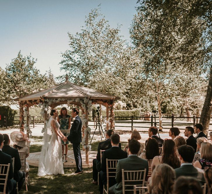 Outdoor Home Wedding Ceremony in Front of Pergola with Bride in Pronovias Danaia Wedding Dress and Groom in Navy Moss Bros. Suit