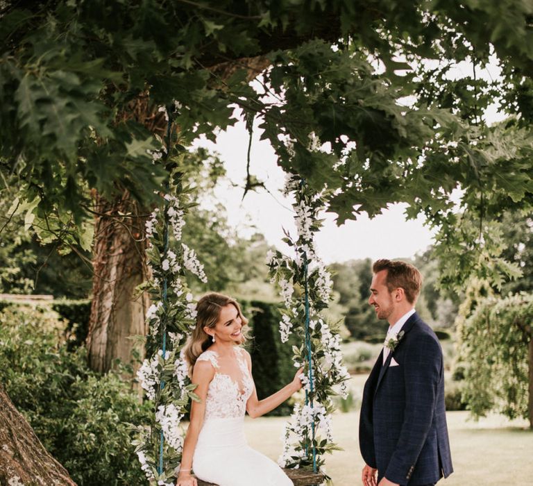 Bride in Pronovias Wedding Dress Sitting on a Ivy Covered Swing with Groom in Moss Bros. Suit  Standing By