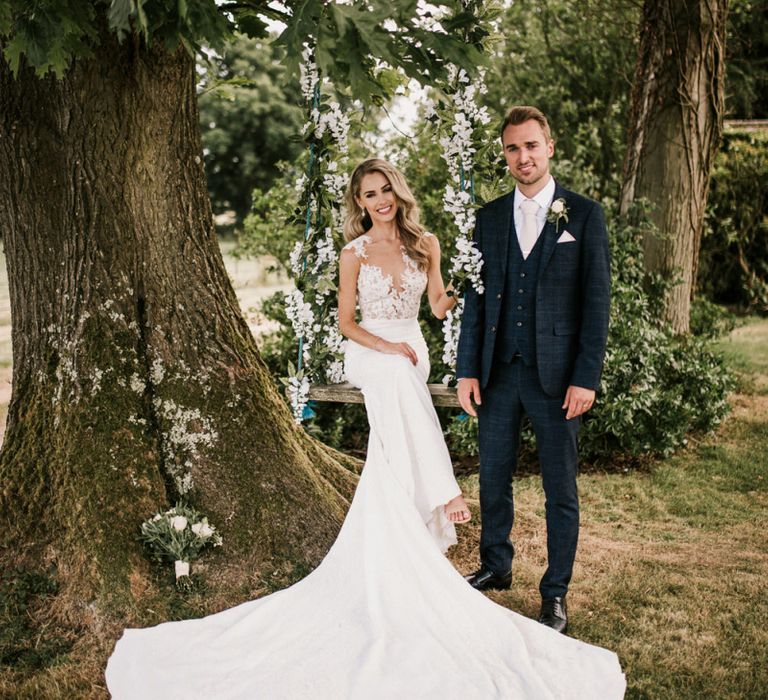 Bride in Pronovias Wedding Dress Sitting on a Ivy Covered Swing with Groom in Moss Bros. Suit  Standing By