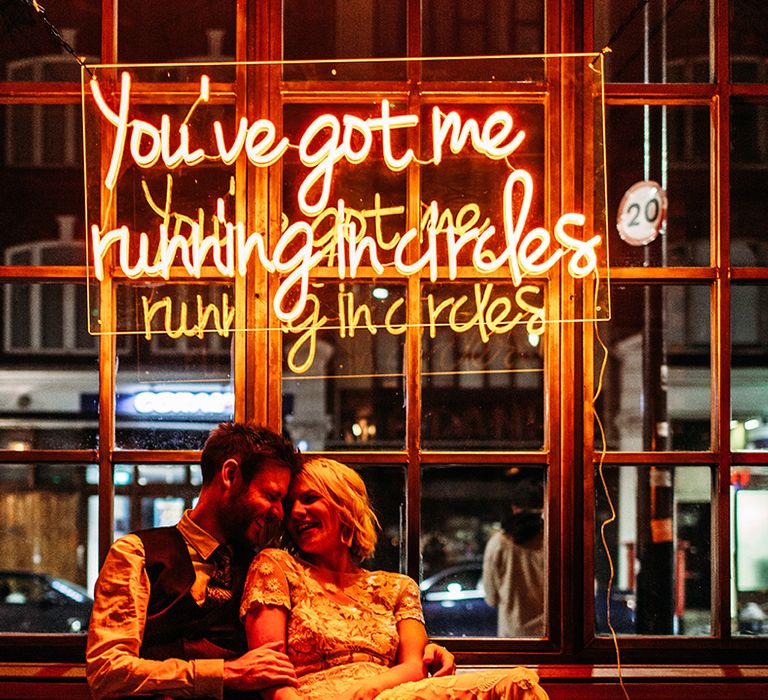 Bride in Lace Hermione De Paula Wedding Dress and Groom in Check Ted Baker Suit Sitting Under Bespoke Neon Sign by Bag and Bones