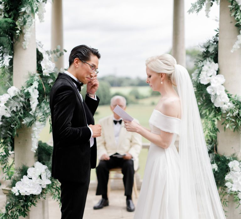 Bride in off the shoulder wedding dress and groom in tuxedo exchanging vows