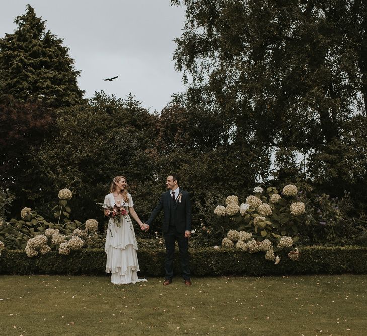 Bride in Tiered Houghton NYC Wedding Dress and Groom in Check House of Fraser Suit Holding Hands in the Garden