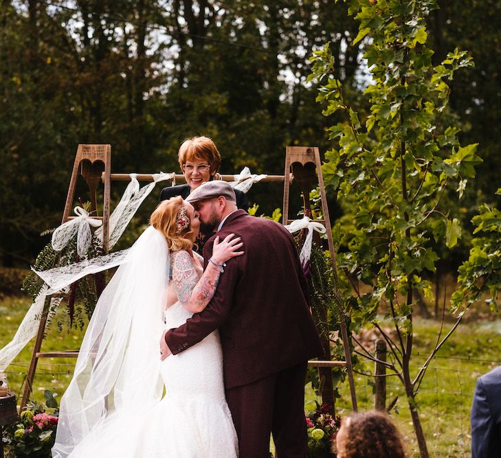 Wedding Ceremony | First Kiss | Bride in Strapless Sassi Holford Wedding Dress with Sweetheart Neckline | Groom in Burgundy Tweed Three Piece Suit | Edison Bulb Floral Installation at Kingsthorpe Lodge Barn Wedding | Johnny Dent Photography