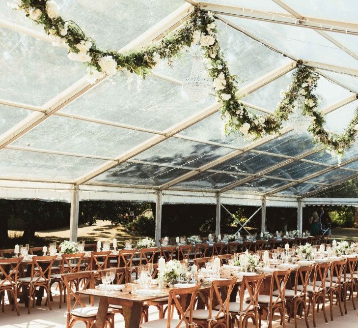 Glass Marquee | Hanging Garland with White Flowers and Greenery | Long Wooden Farm Tables | Wooden Cross Back Chairs | Glass Charger Plates | Gold Cutlery | Gold Rimmed Glassware | White Pillar Candles | Glass Candlesticks | Quintessential English Country Wedding in Glass Marquee at Family Home | Maryanne Weddings Photography