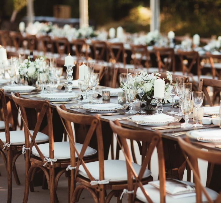 Long Wooden Farm Tables | Wooden Cross Back Chairs | Glass Charger Plates | Gold Cutlery | Gold Rimmed Glassware | White Pillar Candles | Glass Candlesticks | Table Arrangements of White Flowers and Greenery | Quintessential English Country Wedding in Glass Marquee at Family Home | Maryanne Weddings Photography