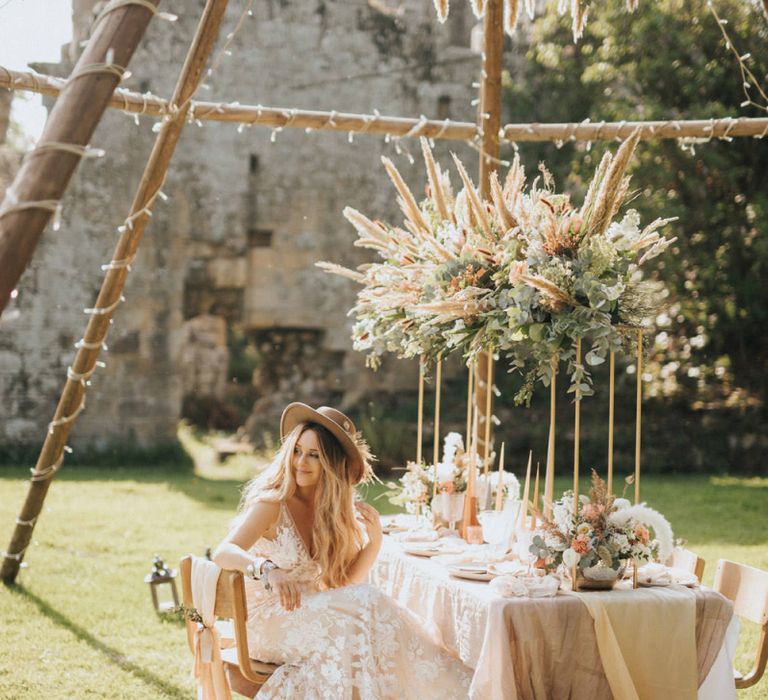 Boho Bride with Straw Hat Sitting at a Tablescape  with Floral Installation Under a Naked Tipi