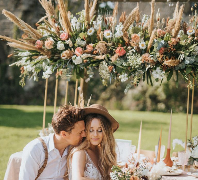 Boho Bride in Lace Wedding Dress and Fedora Hat and Groom in Braces Sitting at Outdoor Tablescape