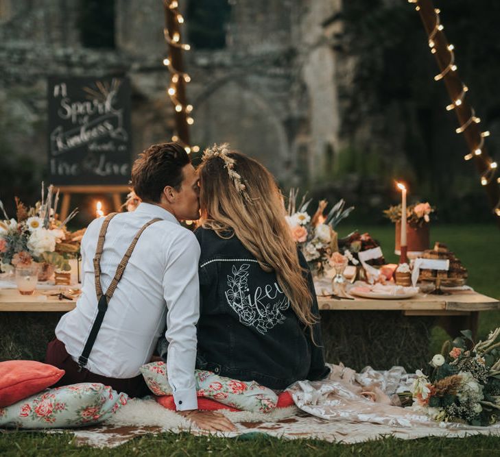 Boho Bride in Customised Leather Jacket and Groom in Braces Kissing at Candlelit Dessert Table