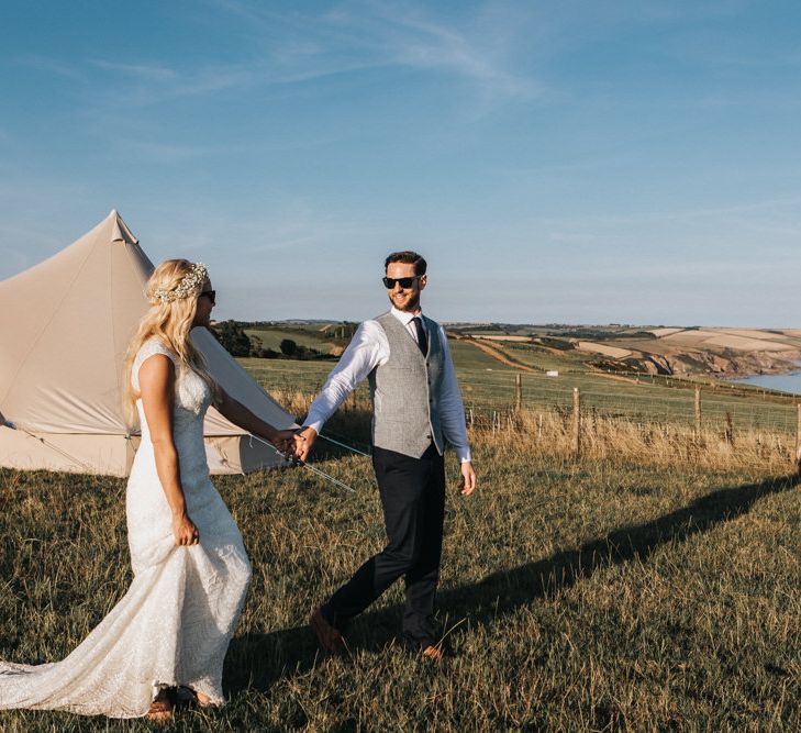 Bride in Sophia Tolli Wedding Dress and Groom in Grey Waistcoat Walking on Cliff Where their Bell Tent is Pitched