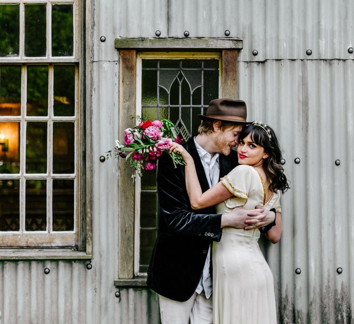 Groom in Navy Velvet Blazer and Hat Hugging His Beautiful Bride in Vintage Wedding Dress with Gold Trim-110