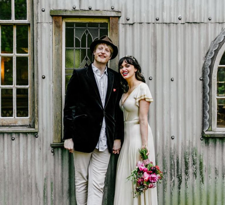 Bride in Vintage Wedding Dress with Gold Trim and Groom in Navy Velvet Blazer and Hat Embracing