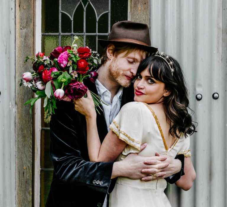 Bride in Vintage Wedding Dress with Gold Trim and Groom in Navy Velvet Blazer and Hat Embracing