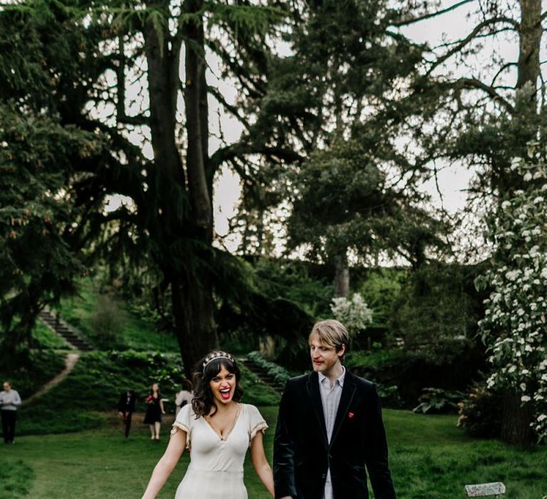 Bride in Vintage Wedding Dress with Gold Trim and Groom in Navy Velvet Blazer and Hat Waking Hand in Hand-114