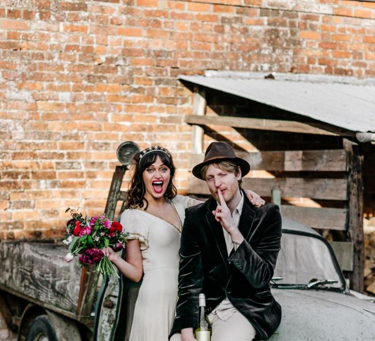 Bride in Vintage Wedding Dress with Gold Trim and Groom in Navy Velvet Blazer and Hat Sitting on an Old Truck-115