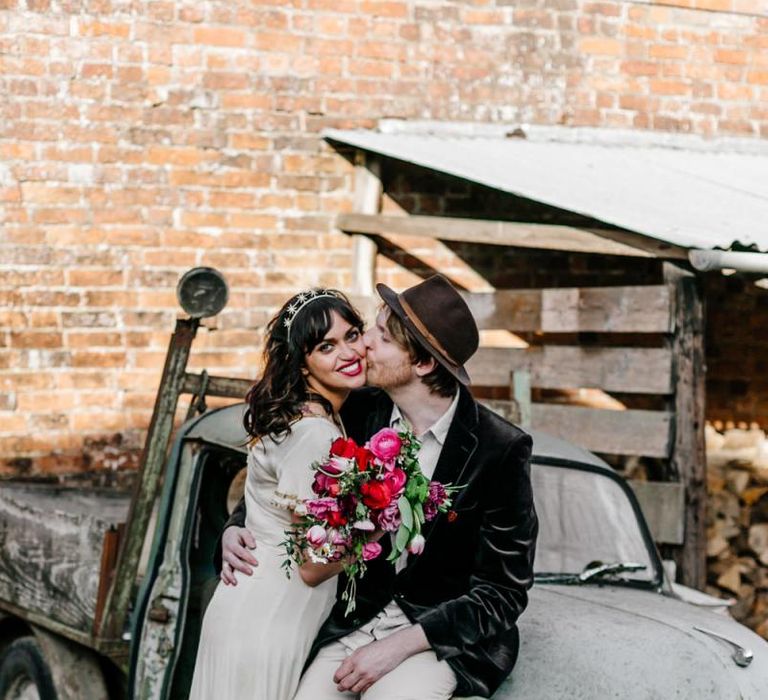 Bride in Vintage Wedding Dress with Gold Trim and Groom in Navy Velvet Blazer and Hat Sitting on an Old Truck Kissing