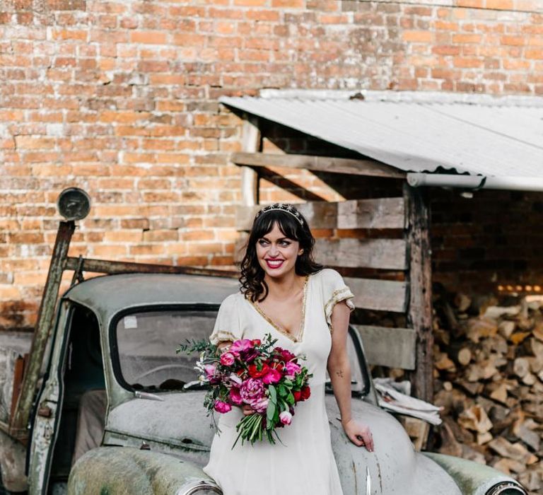 Bride in Vintage Wedding Dress with Gold Trim Holding a Red and Pink Wedding Bouquet Sitting on an Old Truck