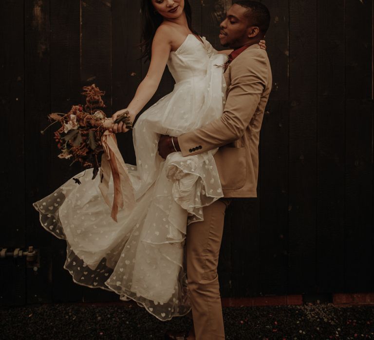 Groom in beige wedding suit picking up his bride in an embroidered wedding dress at the Giraffe Shed