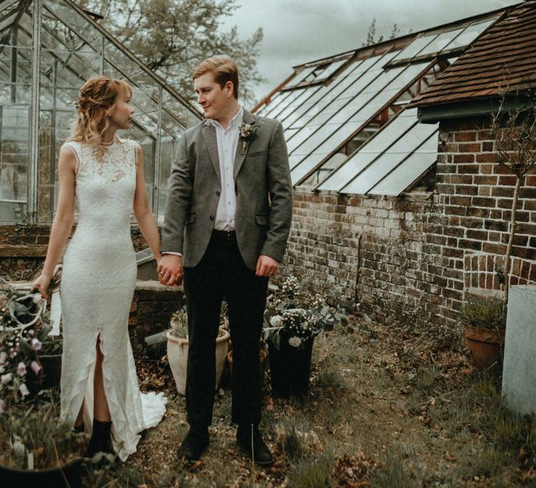 Bride in Lace Wedding Dress with Front Split and Groom in Wool Blazer Holding Hands by a Greenhouse