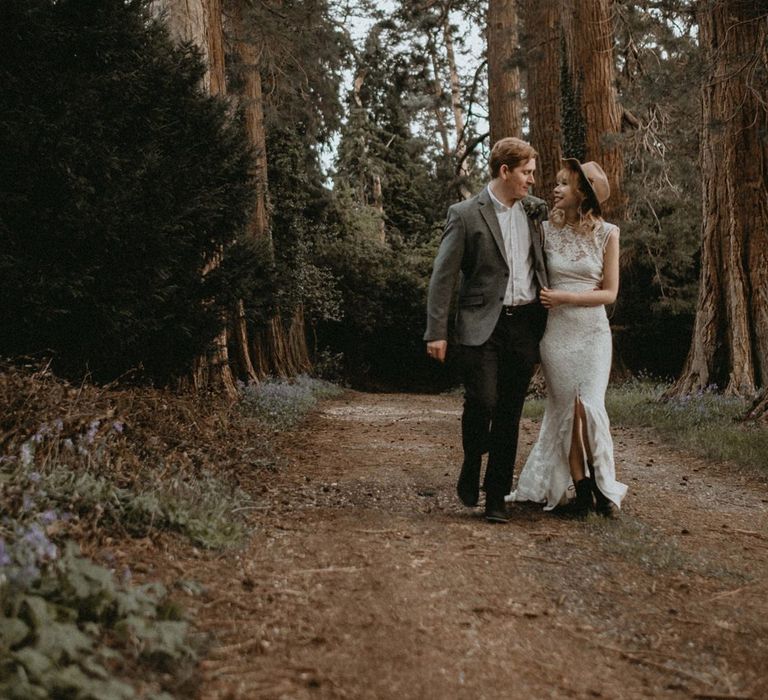 Bride in Lace Wedding Dress and Felt Hat and Groom in Wool Blazer Walking Through The Woodlands