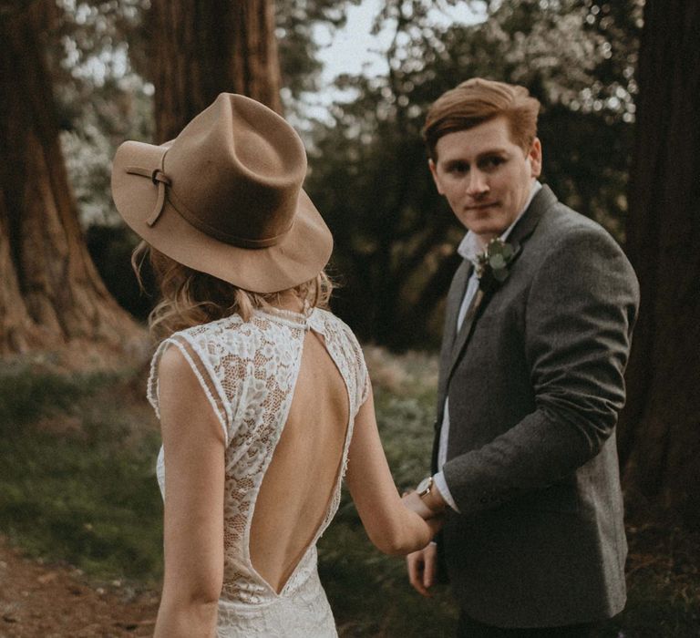 Bride in Backless Lace Wedding Dress and Felt Hat and Groom in Wool Blazer Holding Hands Walking Through The Woods