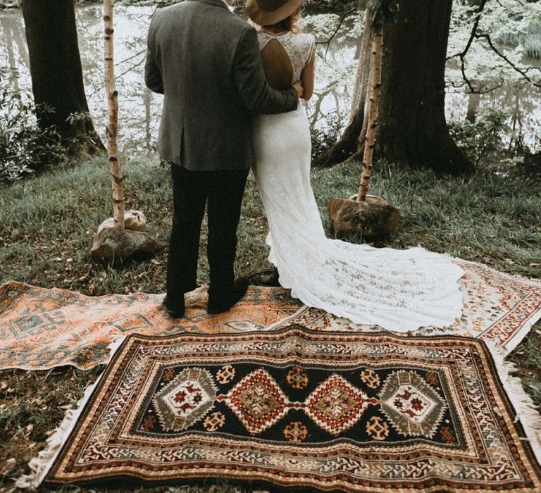 Boho Bride in Lace Wedding Dress and Felt Hat and Groom in Wool Blazer  Standing in Front of Wooden Frame and Rug Altar