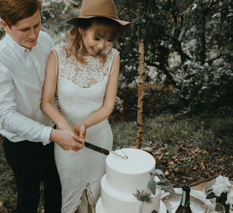 Boho Bride in Lace Wedding Dress and Felt Hat Cutting the White Buttercream Wedding Cake