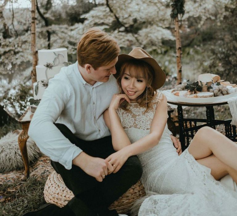 Boho Bride in Lace Wedding Dress and Felt Hat and Groom in White Shirt  Embracing on Chill Out Area