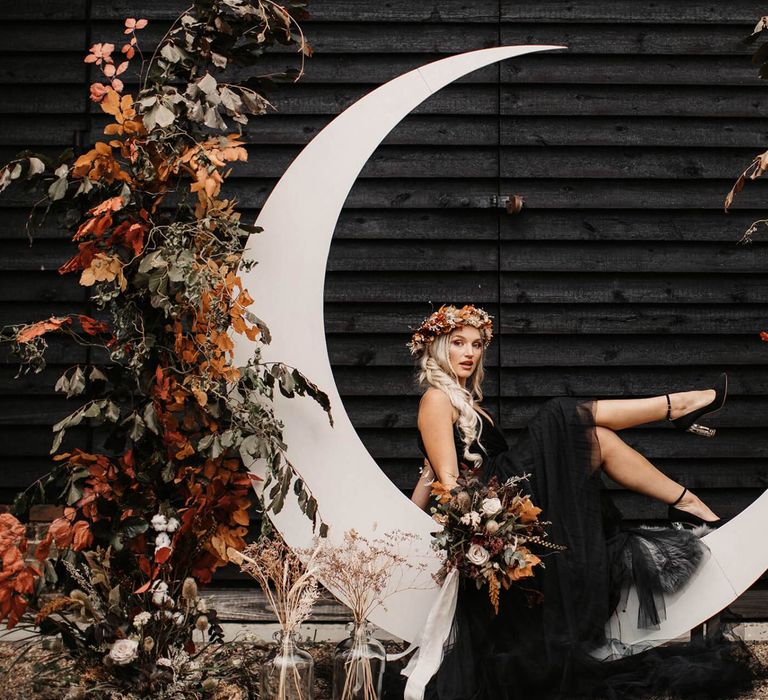 Stylish bride in black wedding dress sitting on a crescent moon  at Halloween wedding