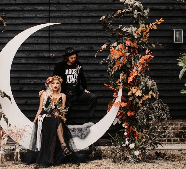 Stylish bride and groom sitting on a crescent moon flanked by autumnal floral arrangements  at Halloween wedding