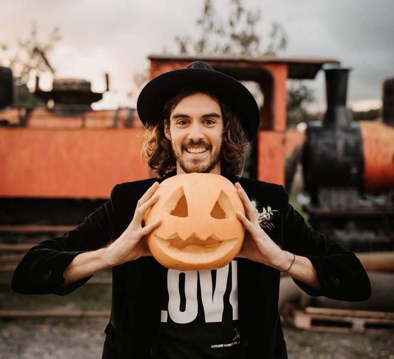 Groom in t-shirt, blazer and hat holding at carved pumpkin at Halloween wedding