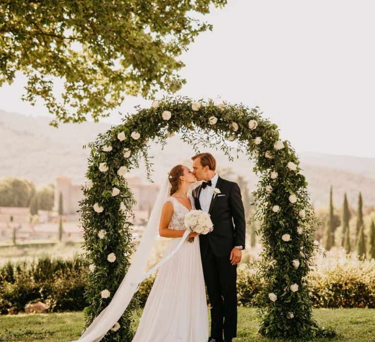 Bride in Bespoke Wedding Dress and Groom in Tuxedo Kissing in Front of White and Green Floral Arch