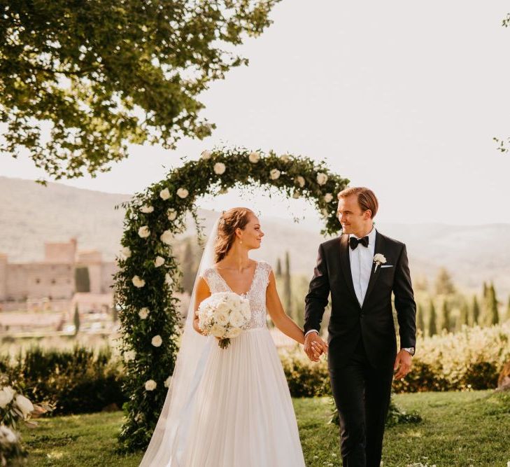 Bride in Bespoke Wedding Dress and Groom in Tuxedo Holding Hands in Front of White and Green Floral Arch